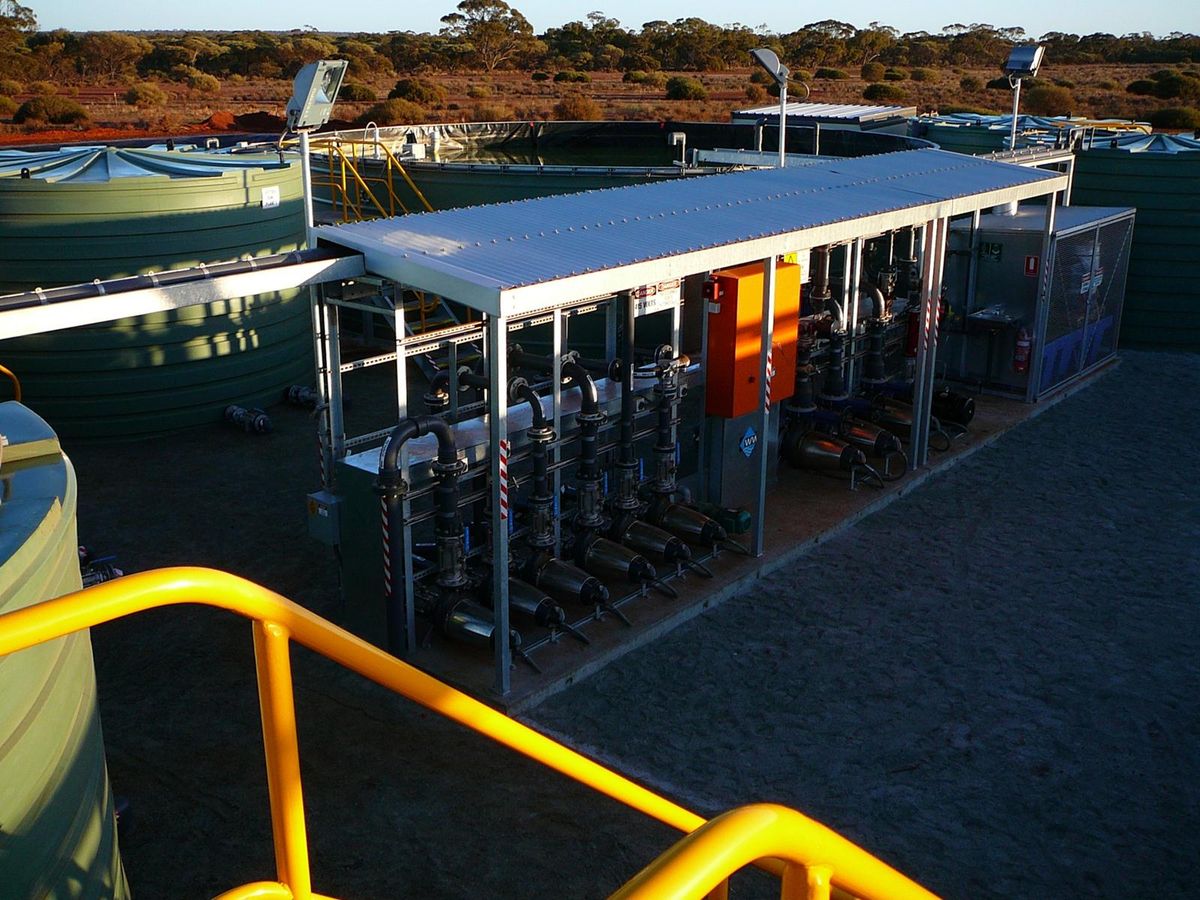 A roofed plant module seen from above, a row of pumps and valves along its length, poly tanks alongside.