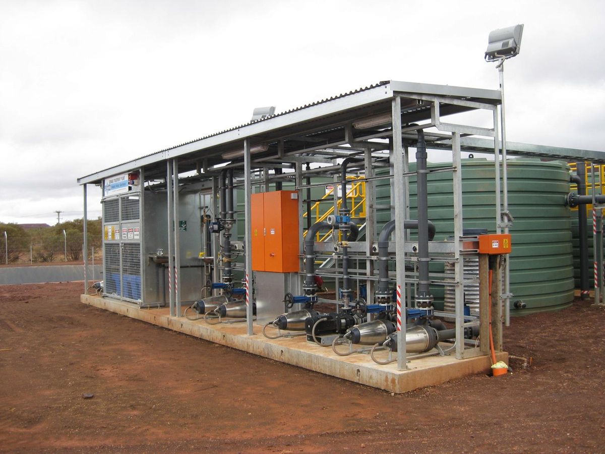 A roofed equipment deck at a treatment plant: switchboards, valves, pipework and pumps on a concrete slab beside poly storage tanks.